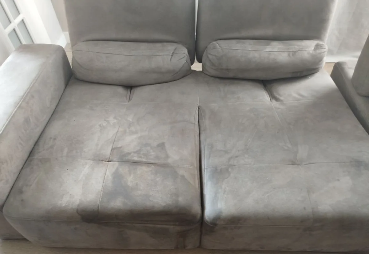The same grey suede sofa before cleaning &mdash; dark greasy patches and watermarks on the seats.